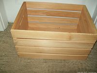 Side angled view of the unfinished wooden crate showing horizontal slats and cutout handle on side.