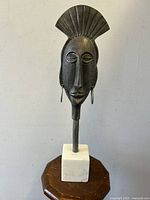 Front view of decorative elongated female head metal sculpture with fan headdress and marble base, showing hoop earrings and stylized features.