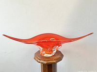 Wide frontal view of the red-orange winged glass bowl on a small round wooden pedestal, showcasing the vibrant color and winged shape.
