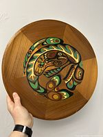 Top view of round wooden plaque with painted raven and whale Indigenous design, held in hand to show size and details.
