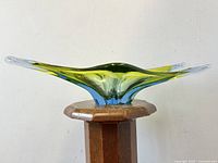 Front view of asymmetrical glass bowl with blue and green tint on wooden stand, showing shape and color gradient.