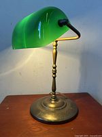 Front angle of the vintage banker’s table lamp, showing lit green Emeralite glass shade and brass-tone base.