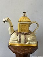 Side view of ceramic camel and rider novelty teapot on wooden surface, showing the full body of the camel and the rider-shaped lid.