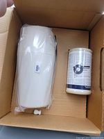 Photo of white plastic water filter housing and cylindrical filter cartridge inside a cardboard box.