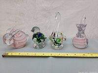 Set of four colourful glass animal figurines arranged with a tape measure for size reference, featuring the fish, duck, swan and cat-like figure.