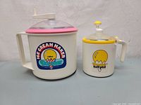 Photo showing both manual ice cream makers side by side with clear lids and colored rims, one large and one half-pint size.