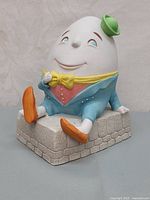 Front-left angle view of Humpty Dumpty figurine seated on grey stone base showing detailed face and colorful outfit.