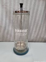 Full front view of clear glass Barbicide jar with white lettering, metal lid, black lid knob, and blue base ring.