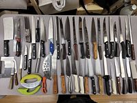 Top-down view of a wide variety of kitchen knives with different handle colors and blade types, alongside cutting utensils like peelers and mandoline slicers.