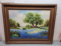 Full view of the framed oil painting showing a scenic landscape with a large tree and field of blue flowers.
