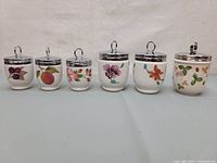 Full view of six Royal Worcester porcelain egg coddlers with floral and fruit designs and chrome lids.