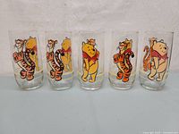 Set of five clear glass tumblers decorated with colorful Disney characters Pooh and Tigger on a plain surface.