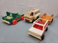 Four vintage Fisher-Price Adventure People vehicles from 1974 on plain grey surface showing green Jeep, white police van, white/red SUV, and yellow trailer.
