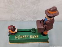 Front side view showing the monkey on the left and man with suitcase on the right on green base with 'MONKEY BANK' lettering.