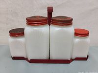 Front view of four milk glass containers in red metal holder with red metal lids, showing set from one angle.