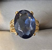 Front view of gold plated ring with large blue faceted stone showing oval shape and prong setting.