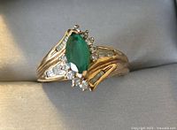 Front angled view showing marquise green stone surrounded by round clear stones and baguette clear stones along gold plated band.