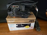 Photo showing Skil Classic Series 3 inch x 18 inch belt sander in black with power cord, sitting on original box, dust bag rolled beside it.