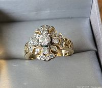 Close-up image showing the top face of a gold-tone fashion ring with clear round gemstones arranged in a cross/flower pattern. Light shines on facets of the stones, all mounted on a gold-tone band.