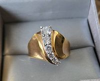 Top-down view of the gold-tone ring showing five clear stones arranged diagonally across the wide band served in a grey jewelry box.