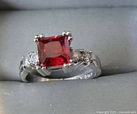 Close-up photo of fashion ring showing square red central stone and clear side stones on silver-tone band.
