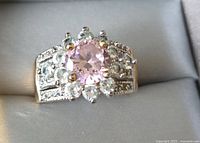 Close-up front angled view of round pink center stone surrounded by clear stones on a metal band.