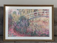 Frontal image of a framed print depicting Claude Monet's painting 'Le Pont Japonais sur Bassin aux Nymphéas'. The print is under glass in a wood frame with decorative edging. The print shows a white curved Japanese bridge over water with pink and green foliage.