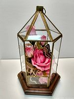 Hexagonal glass display case with brass-colored metal frame on wooden base, containing pink artificial flowers and butterfly ornament.