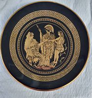 Full view of the circular black plate showing the gold etched Greek figures and the Greek key border pattern with a gold rim.