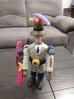 Inspector Gadget plastic figure from 1999 McDonald's Happy Meal, standing on table in front of sofa, showing front side with red and blue gadget attachments on arms and umbrella accessory on hat.