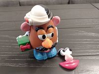 Mr. Potato Head toy with white hat, blue shoes, green bowtie accessory on the side, detachable eyes and lips placed next to it on table.
