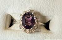 Close-up image of a silver ring with a central oval amethyst gemstone surrounded by a textured silver setting on a white leatherette background.