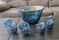Blue carnival glass punch bowl in center with twelve matching cups arranged around it, all placed on a dark table with a brown couch in the background.