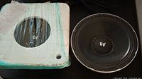 Two Electro Voice EV 12-inch subwoofers shown with one wrapped in foam packaging and another fully visible showing the black cone and EV logo on the dust cap.