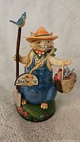 Front view of Jim Shore Heartwood Creek cat figurine with hat, blue overalls, holding staff and basket