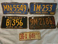 Five vintage license plates shown together: two New York plates, one with yellow on blue MN-5549 and another with 1M 253 and a 1973 sticker, one Arkansas truck plate black with yellow B 356 expiring 6-30-66, one rusty NY Worlds Fair 64 plate 9M 2186, and one small white Nebraska plate with red numbers 36X 648.