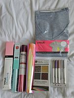 Full lot items displayed on white sheet including T-shirt in packaging, rolled iron-on sheets, Infusible Ink card pack, and five Cricut pens in original backing.