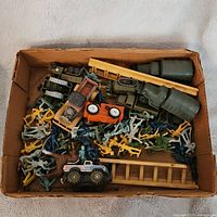 Box view displaying plastic army men, military trucks, wooden ladders, and accessories arranged inside a shallow cardboard box.