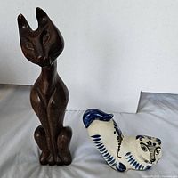 Photo showing both cat figurines, tall wooden carved cat figurine and small ceramic cat figurine with blue decoration.