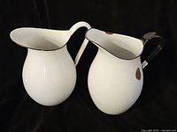 Both white enamelware pitchers with black rims and handles, showing minor chips and wear on enamel surface.