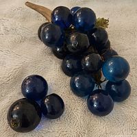 Vintage blue Lucite grape cluster with wooden stem lying on beige fabric. Shows several attached grapes and some loose grapes. Visible wear with three grapes loose from the cluster.