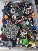 Plastic bin filled with various LEGO pieces including plates, bricks, wheels, and specialty elements in multiple colors.