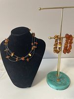 Overall front view of amber jewelry set displayed on black bust and gold-toned stand with green base, showing amber chip bead necklaces and bracelets on thin wire.