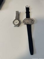 Photo showing two wristwatches: one with metal bracelet and round dial, the other with black leather strap and distorted oval dial with Roman numerals.