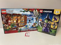 Two LEGO sets displayed side by side: LEGO City Advent Calendar 60235 box with winter cabin and minifigures, and LEGO Creator 3-in-1 Mystic Witch 40562 box featuring a witch minifigure and black cat.
