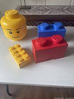 Four LEGO plastic storage bricks on a table, one large yellow head shaped and three brick shaped in red, blue and yellow.