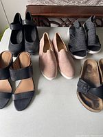 Top view of five pairs of women's shoes in size range 9.5-10 arranged on white surface, black slip-on shoes, pink slip-ons, black fabric wedge shoes, black open-toe sandals, black buckle slides.