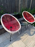 Photo showing two Acapulco-style chairs with red and hot pink cord seats and white frames alongside a glass-top side table on patio stones.