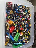 Clear plastic tub filled with multicolored LEGO pieces including bricks, plates, wheels, and specialty parts.