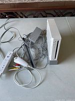 Full view of Wii console standing vertically with cables and accessories spread next to it on table, showing the console, remote, Nunchuk, sensor bar, and cables.